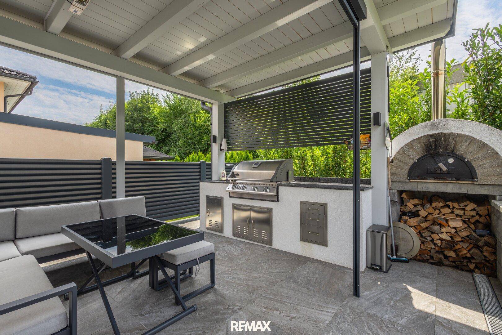 Überdachte Terrasse mit moderner Außenküche, Grill, Pizzaofen und gemütlichem Essbereich im Freien.