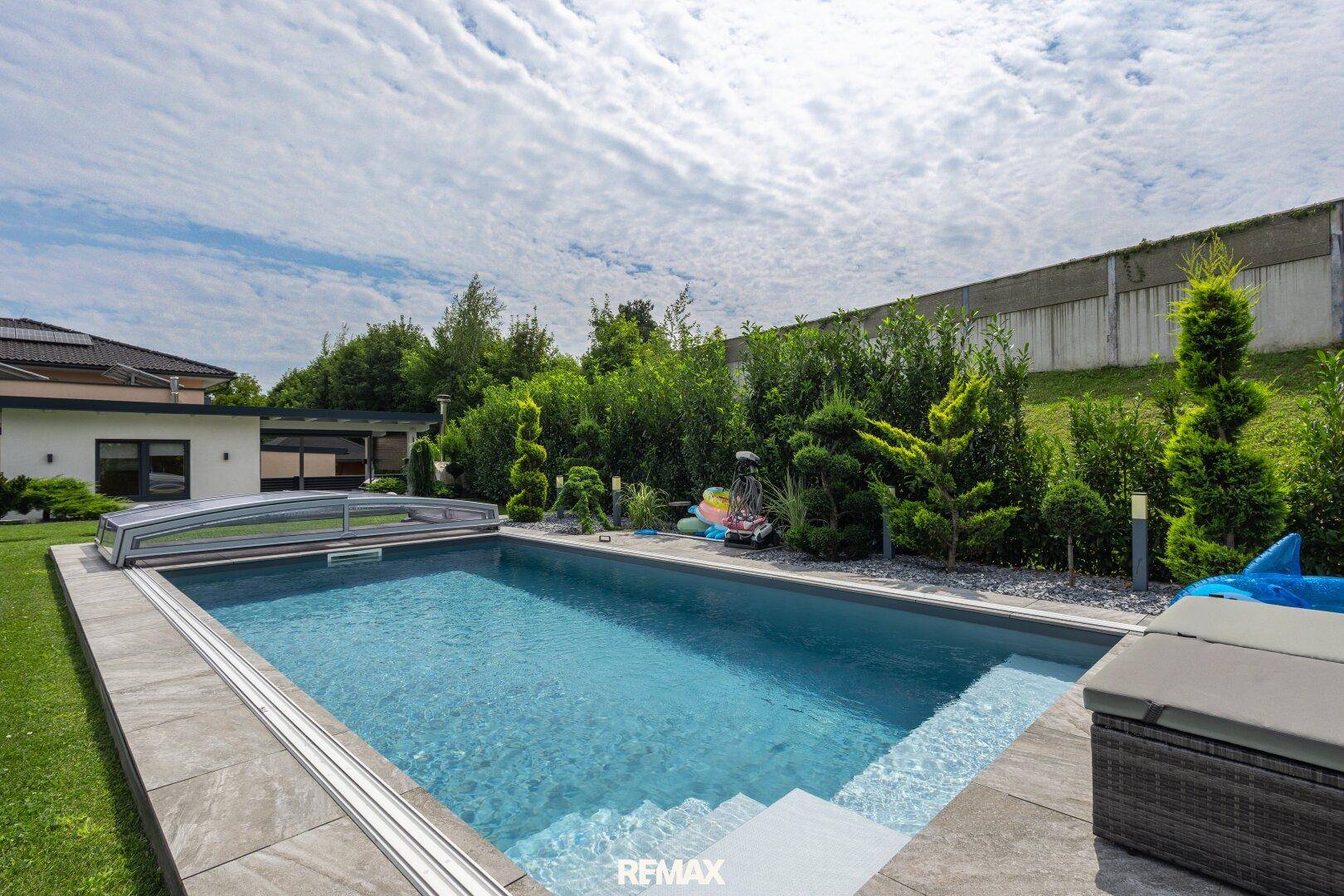 Gepflegter Swimmingpool mit Abdeckung, umgeben von grüner Bepflanzung und einer sonnigen Terrasse.