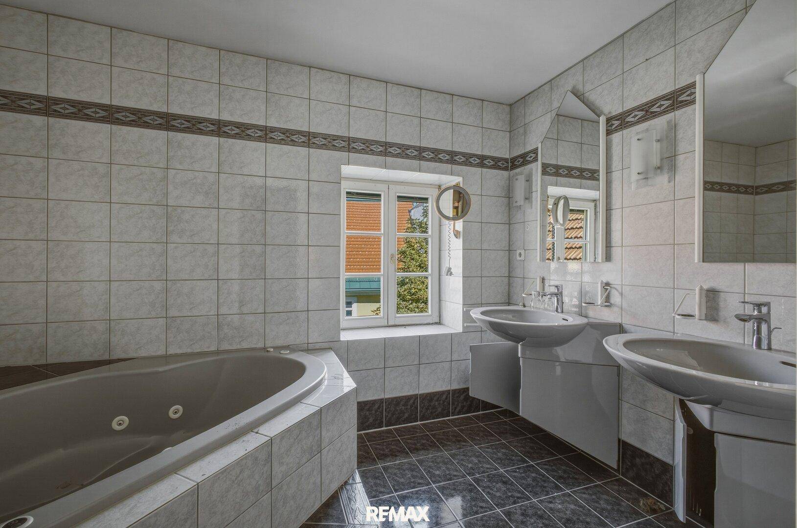 Geräumiges Badezimmer mit Eckbadewanne, Doppelwaschbecken und Fliesen in Hellgrau.