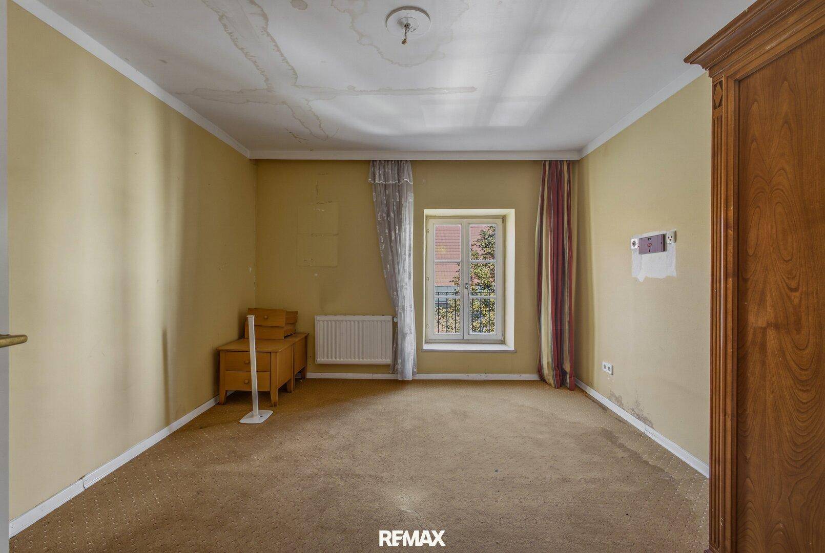 Schlafzimmer mit Teppichboden, gelben Wänden und einem Fenster mit Blick ins Grüne.