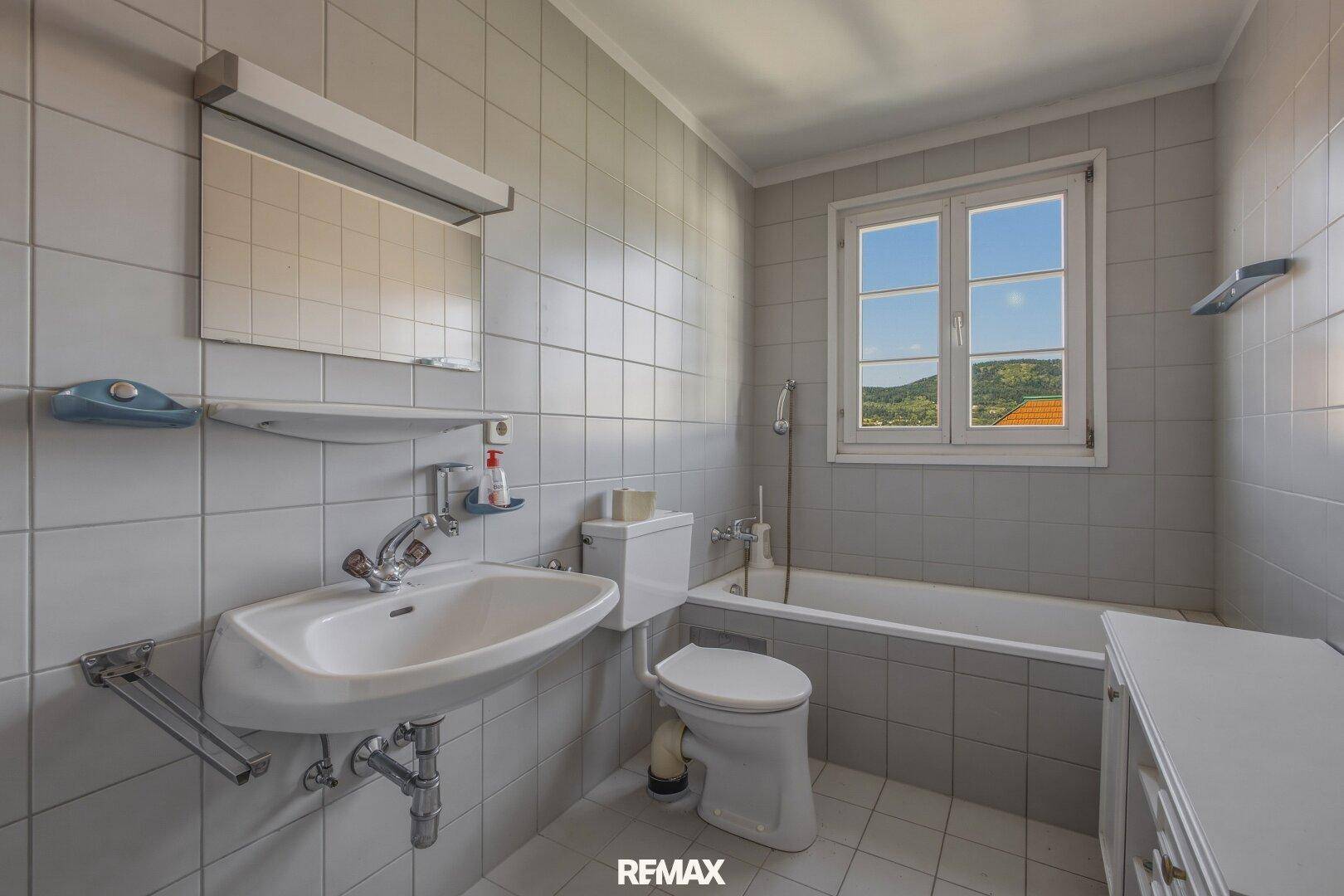 Helles Badezimmer mit Badewanne, WC und Waschbecken, komplett gefliest in Grau.