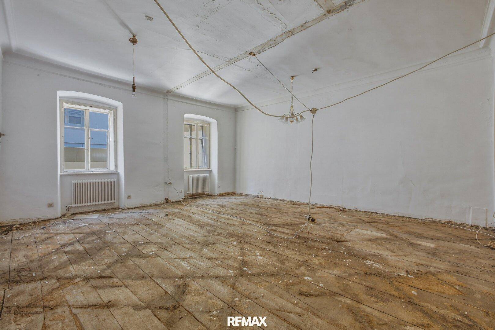 Großer, leerer Raum mit zwei Fenstern und sichtbaren Holzdielen, renovierungsbedürftig.