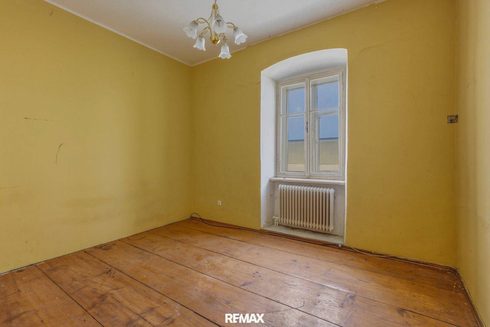 Leerer Raum mit gelben Wänden, Holzdielenboden und einem Fenster mit Heizkörper.