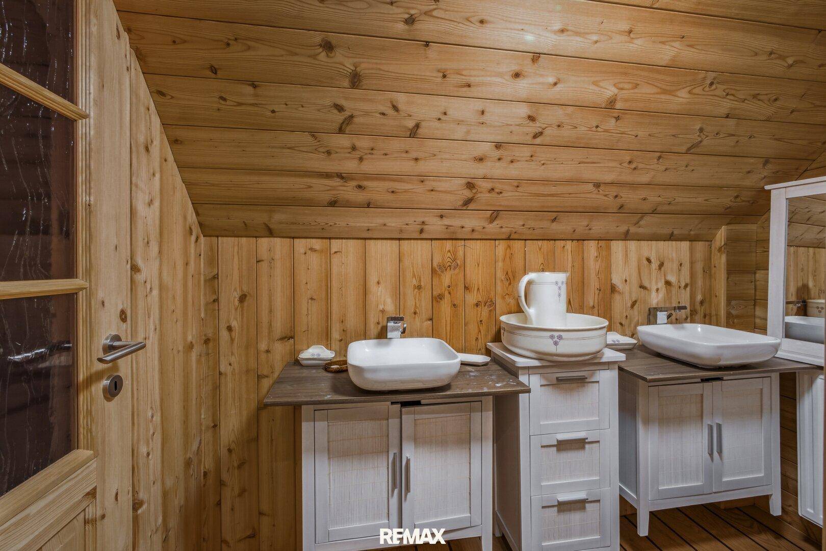 Rustikales Badezimmer mit Holzwänden und Dachschräge, ausgestattet mit zwei Waschbecken und weißen Unterschränken.