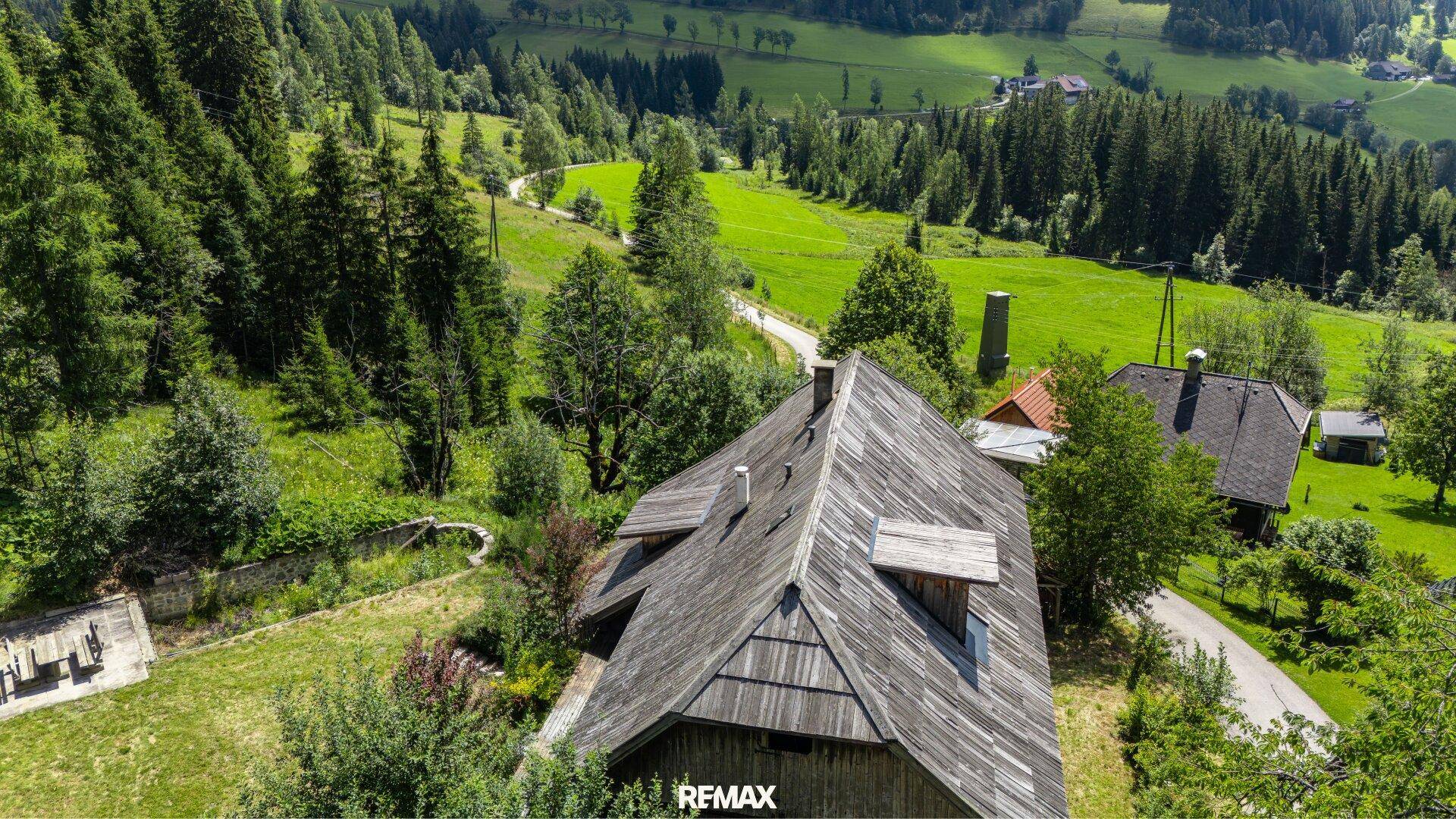 Luftaufnahme eines traditionellen Holzhauses mit Schindeldach, eingebettet in eine grüne Hügellandschaft mit Wäldern.