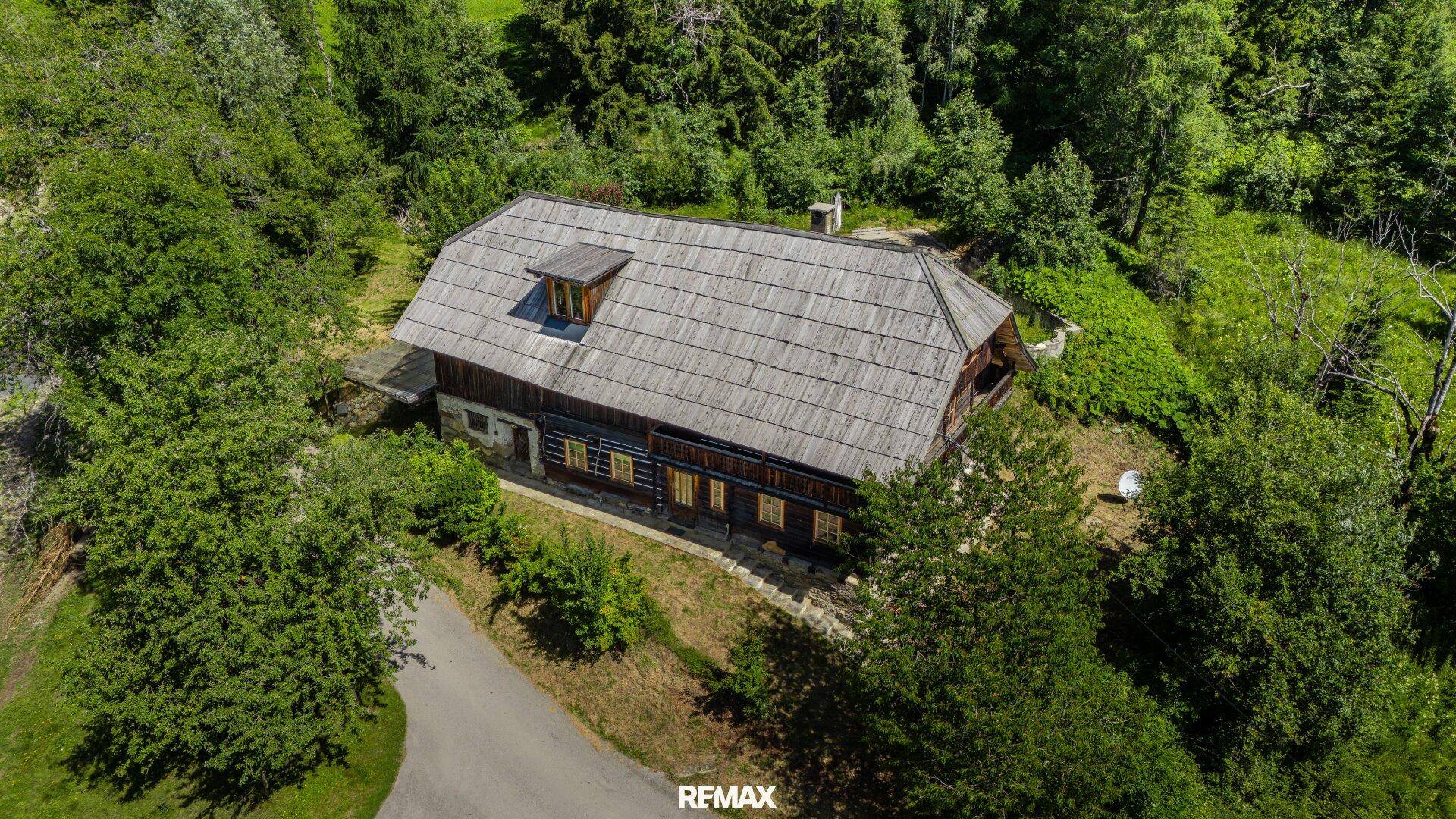 Vogelperspektive eines rustikalen Holzhauses mit Schindeldach, umgeben von dichtem Grün und hohen Bäumen.