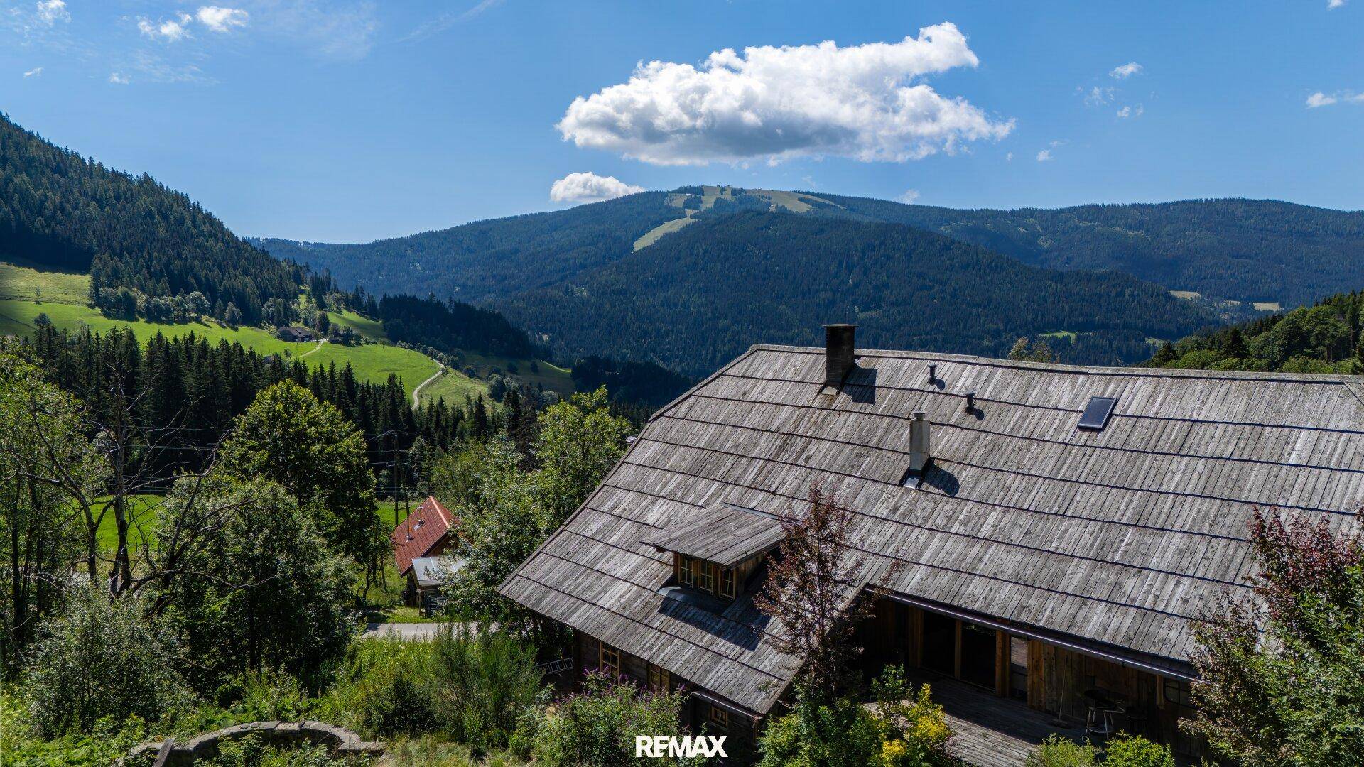 Traditionelles Holzhaus mit Schindeldach und Dachgaube, umgeben von grünen Wäldern und Bergen im Hintergrund.