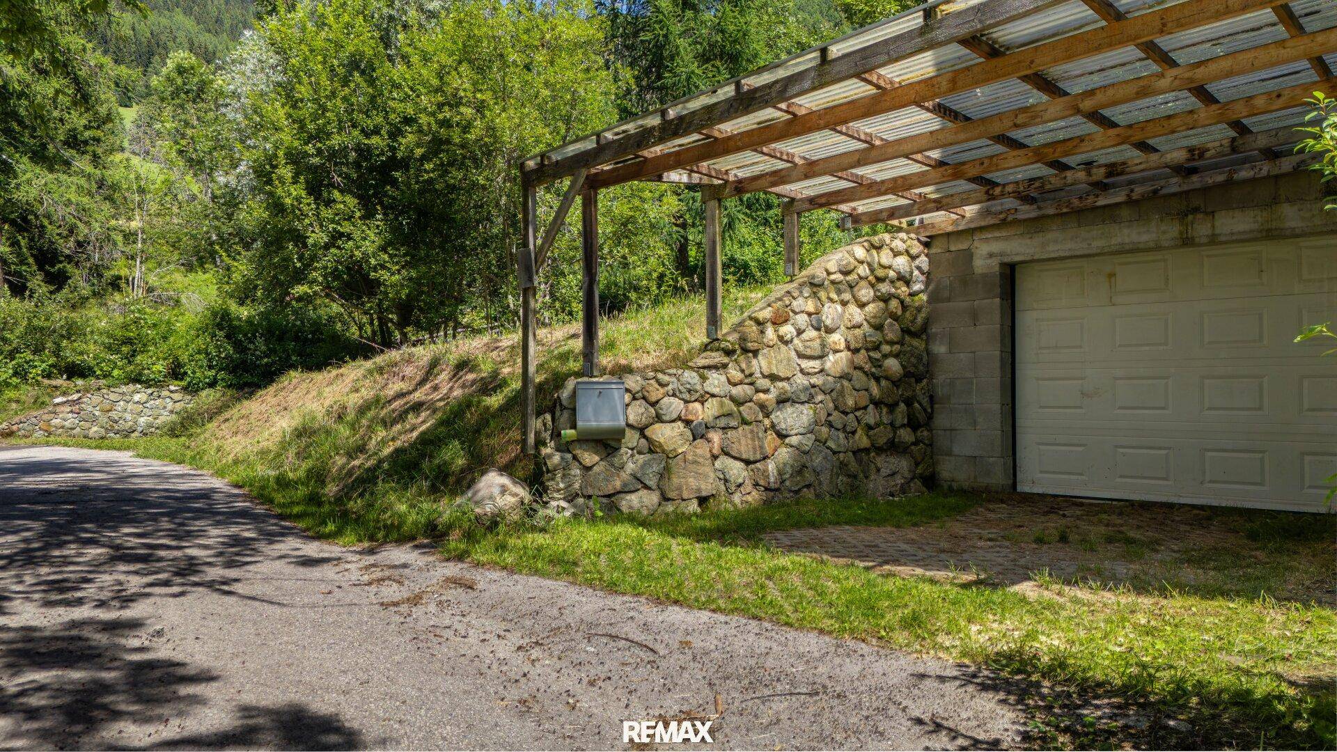 Zufahrt zu einer Garage mit überdachtem Carport und einer rustikalen Steinmauer am Hang.
