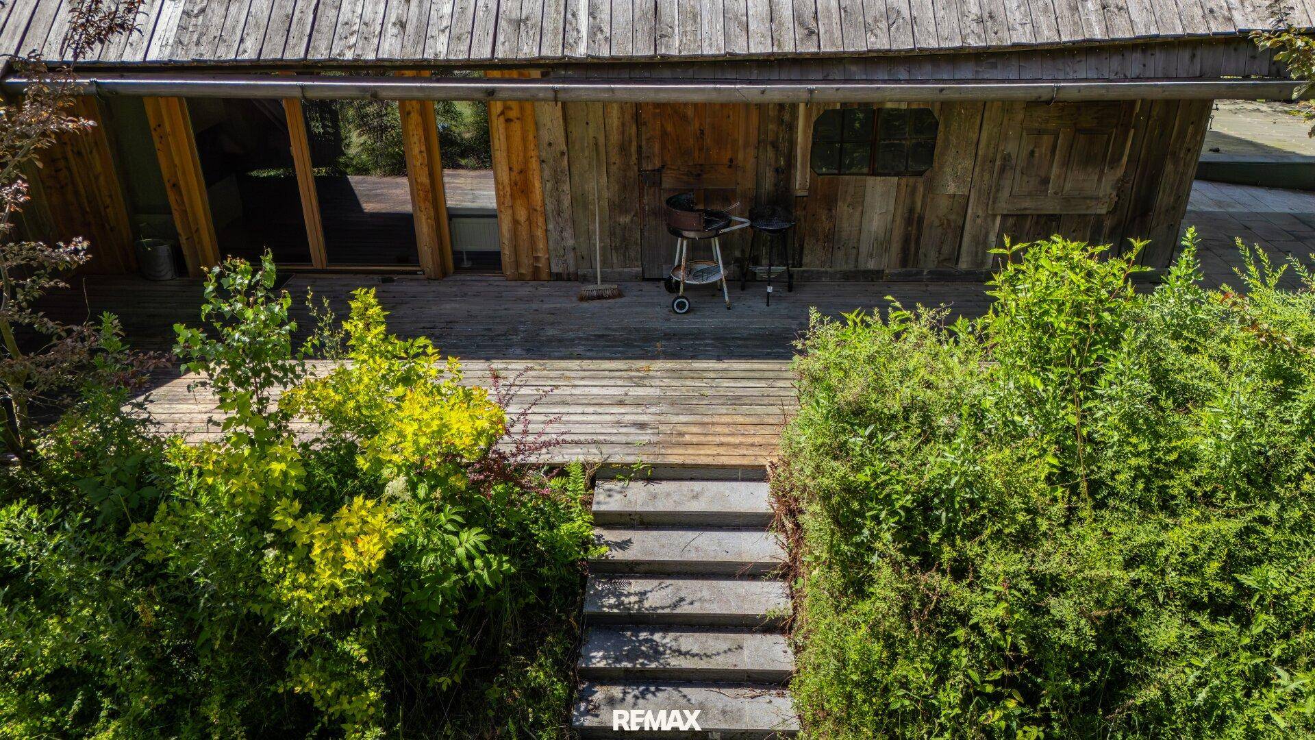 Holzterrasse mit Grillbereich und Zugang zu einem rustikalen Holzgebäude, umgeben von grünen Büschen.