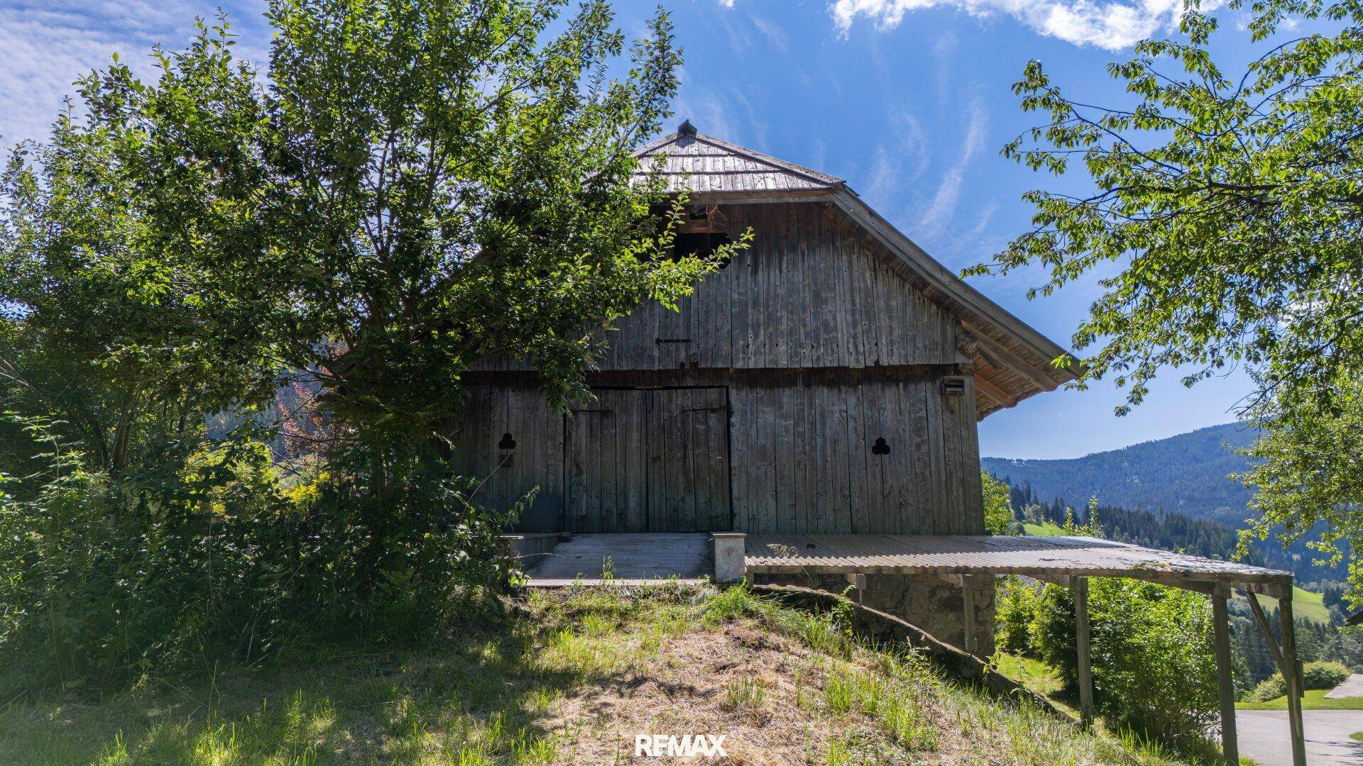 Rustikales Holzgebäude mit Schindeldach und überdachtem Bereich, umgeben von üppiger Vegetation.