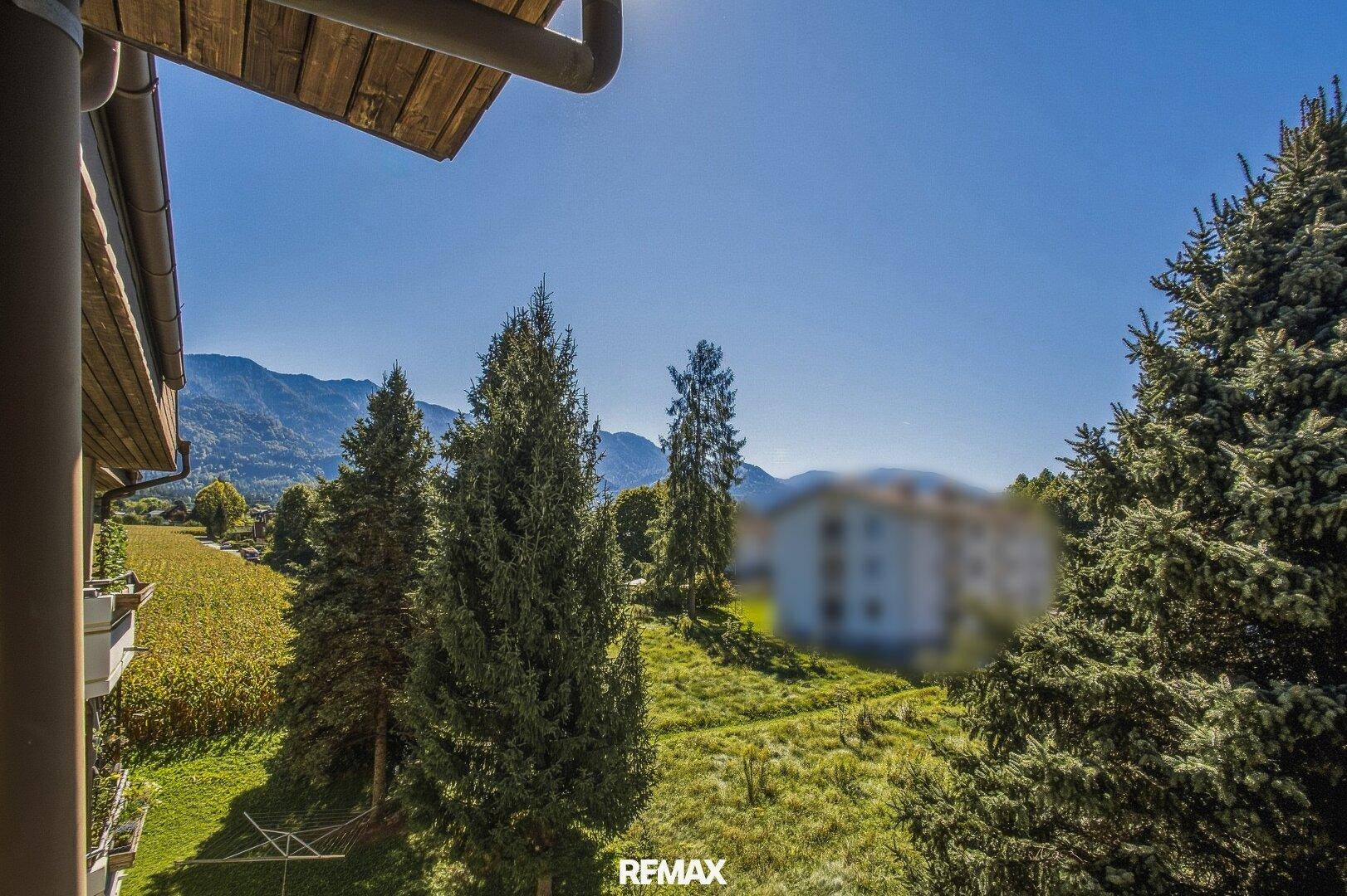 Panoramablick vom Balkon auf grüne Felder, hohe Bäume und die Berge.