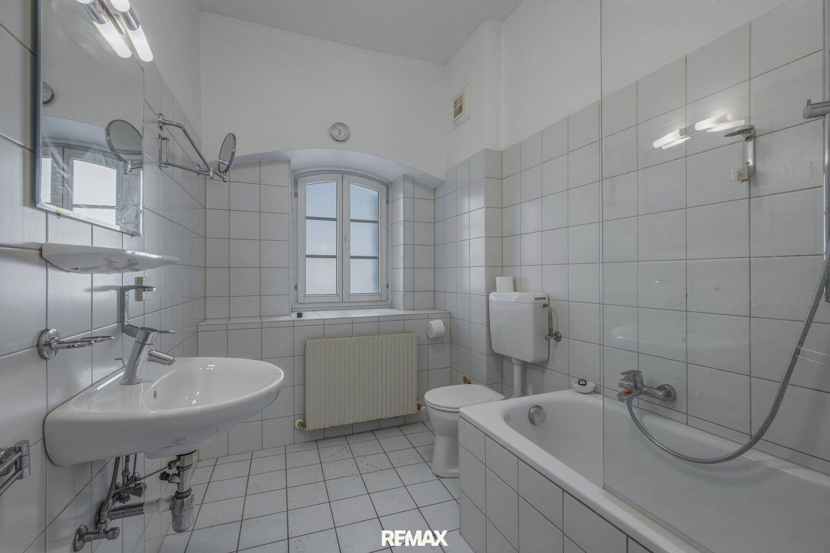 Helles Badezimmer mit Badewanne, Waschbecken und Fenster, gefliest in Weiß.
