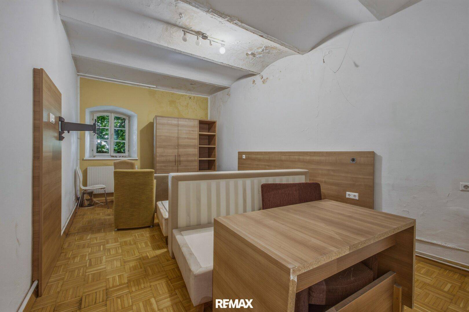 Schlafzimmer mit Bett, Tisch und Kleiderschrank, mit sichtbaren Deckenbalken und Fenster.