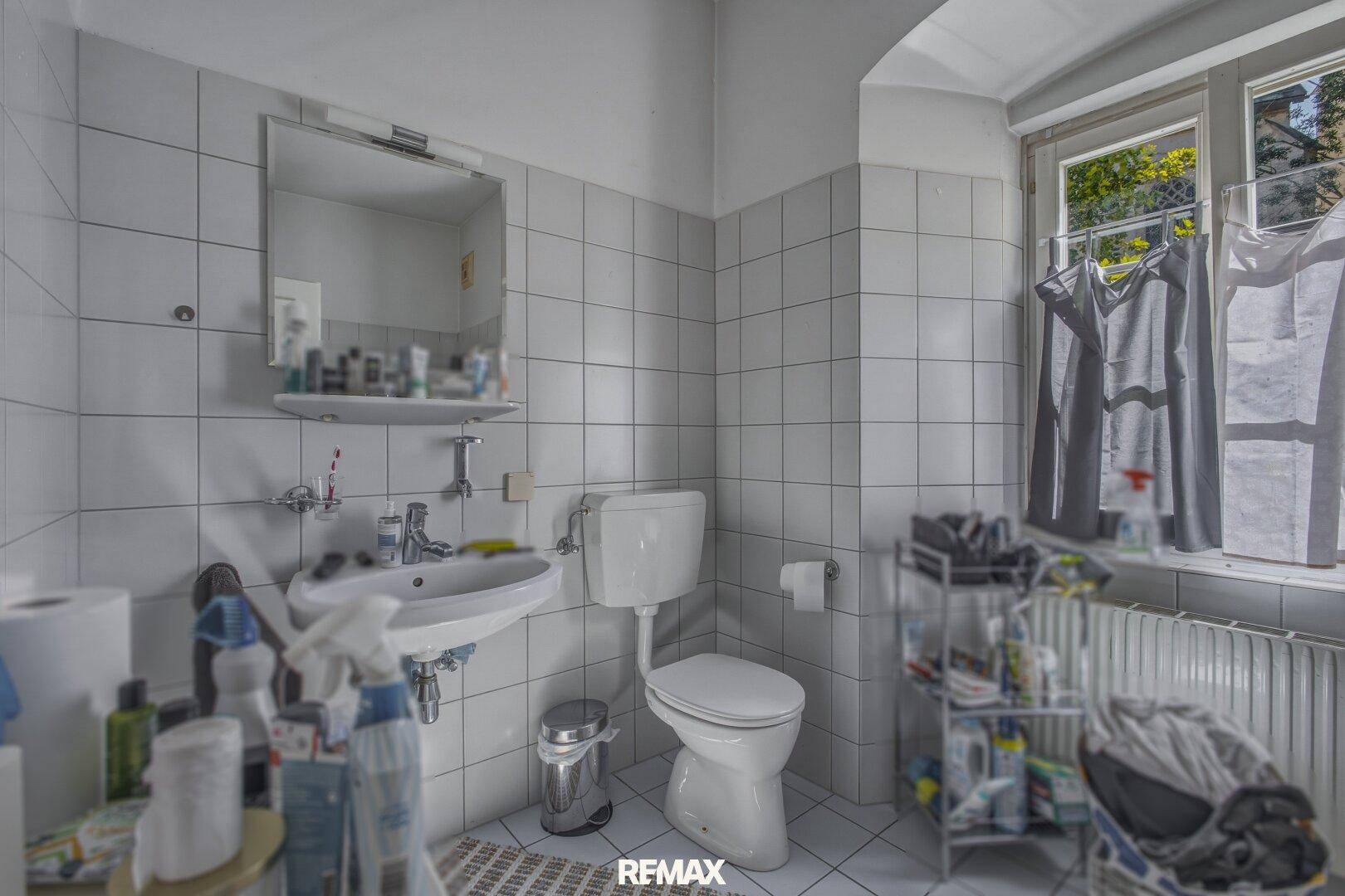 Badezimmer mit Waschbecken, Toilette und Fenster, gefliest in hellem Grau.
