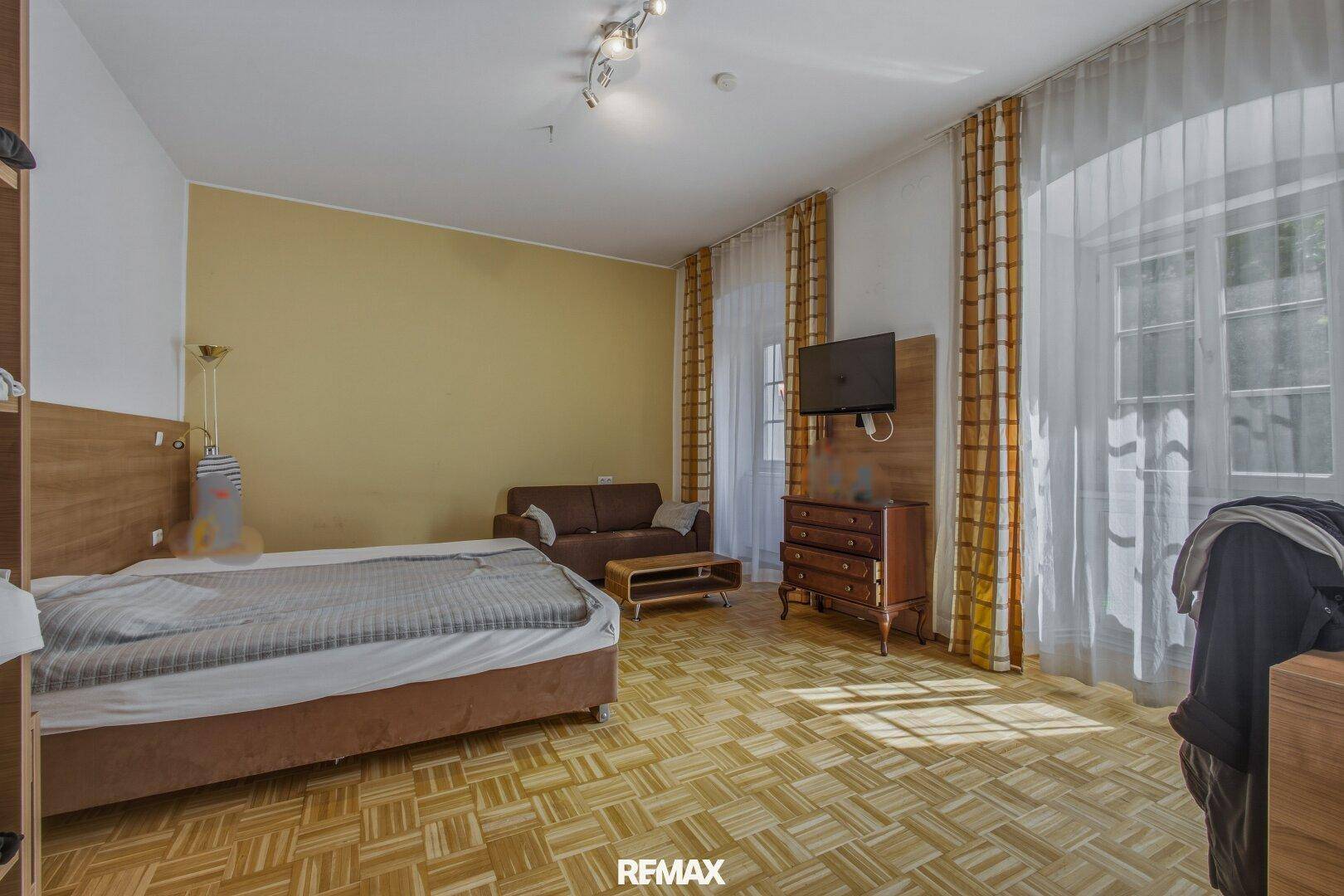 Helles Schlafzimmer mit Doppelbett, Sofa und Fernseher, mit Parkettboden und großen Fenstern.