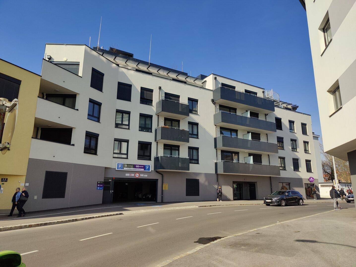 Moderne Wohnanlage mit mehreren Balkonen und Tiefgarageneinfahrt an einer sonnigen Straße.