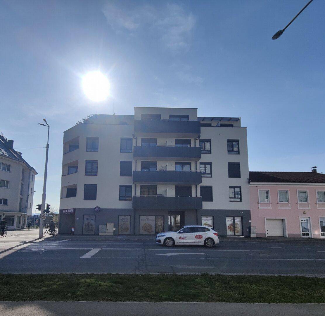 Moderne Wohnanlage mit Geschäften im Erdgeschoss und Balkonen, gegenüber einer belebten Straße.