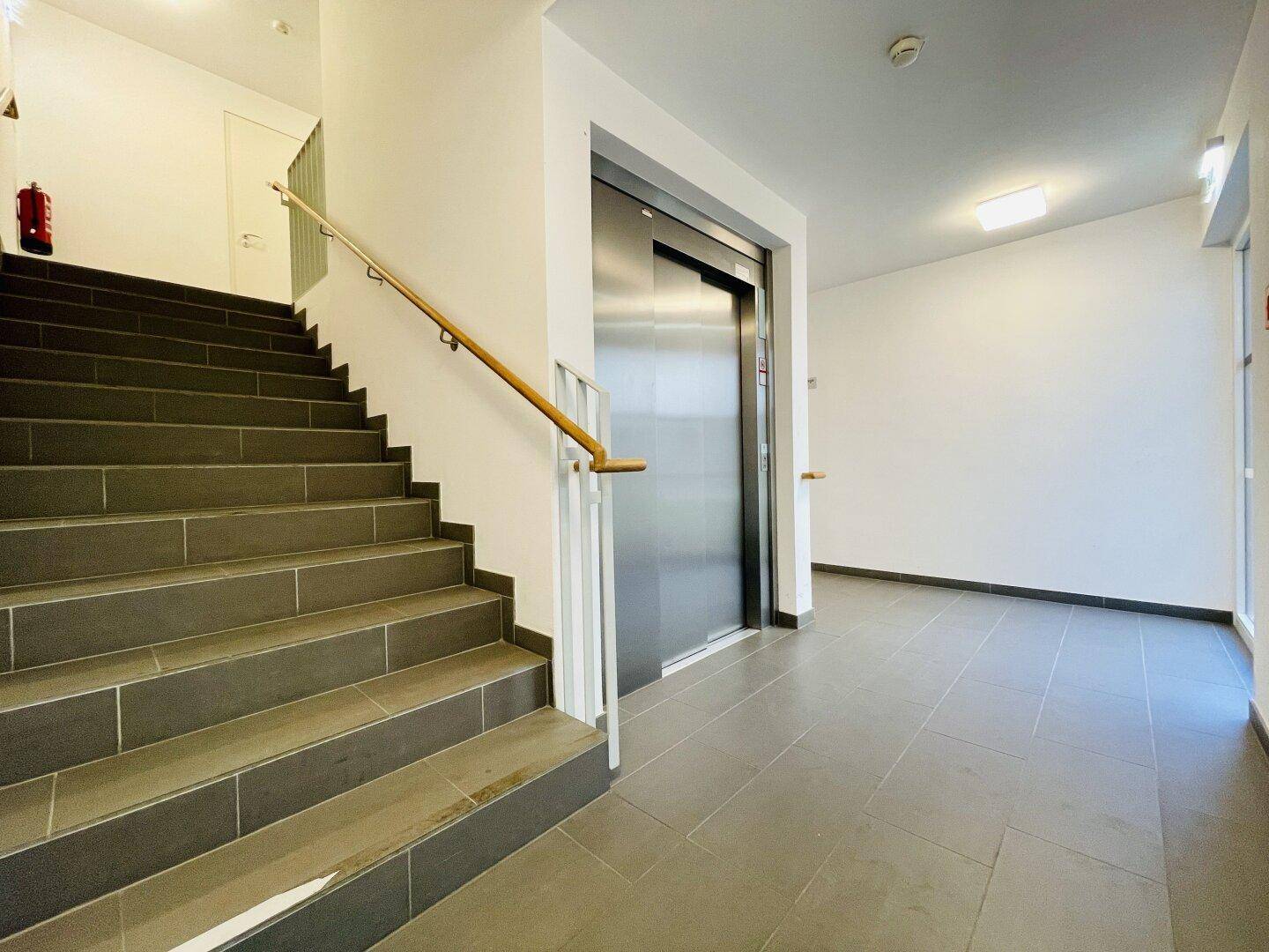Treppenhaus mit Aufzug und Handlauf, ausgestattet mit modernen Fliesen und hellem Licht.