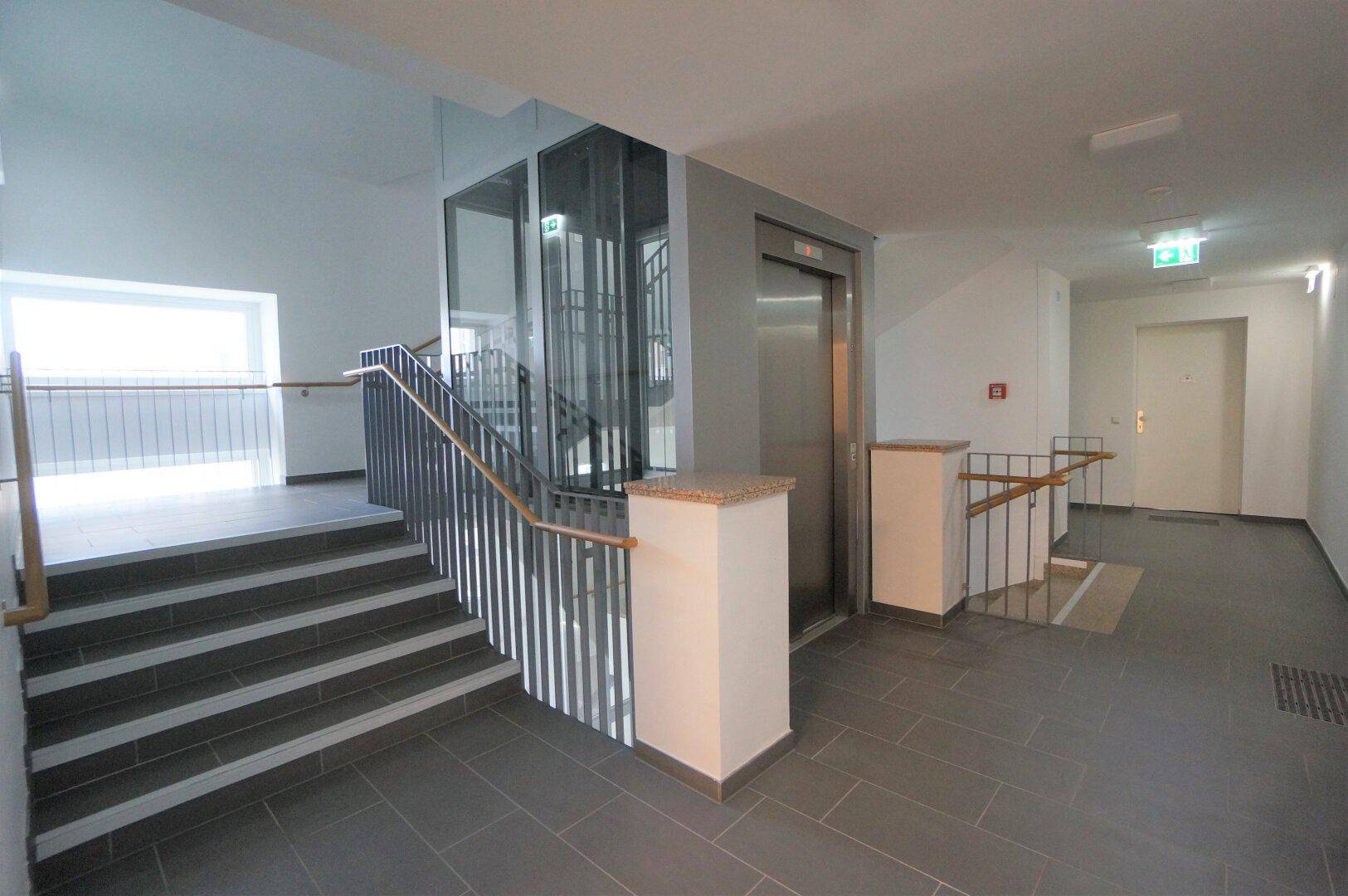 Moderner Eingangsbereich mit Treppe, Glasaufzug und Fliesenboden.