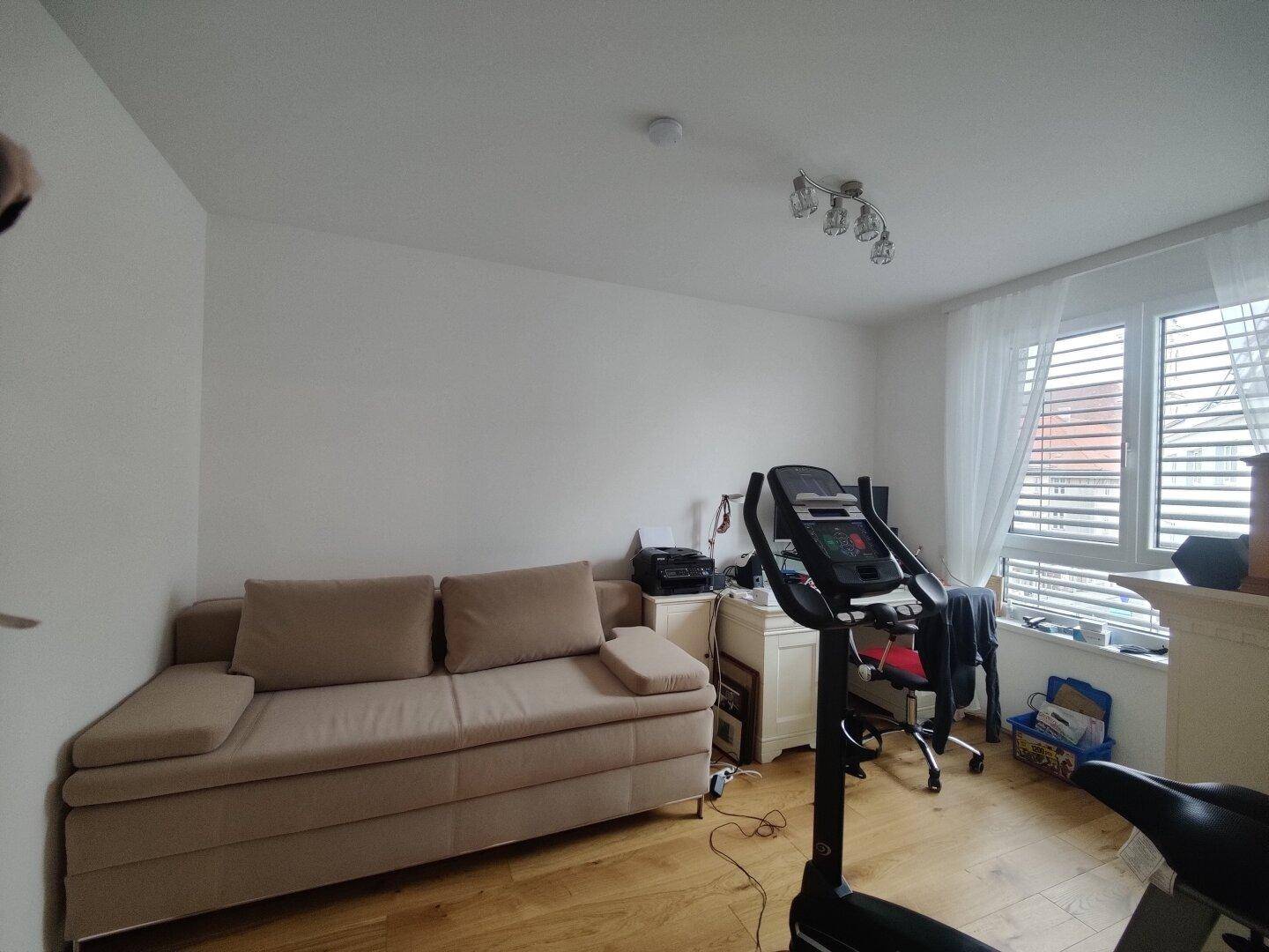 Helles Zimmer mit Schlafsofa, Trainingsgerät und Fenster mit Jalousien.