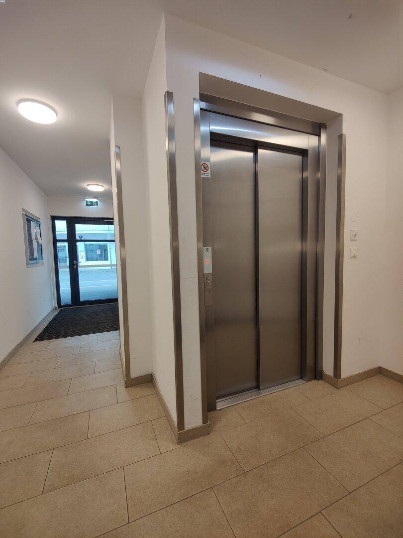 Moderner Aufzug in einem hellen Flur mit Fliesenboden und Zugang zur Straße.
