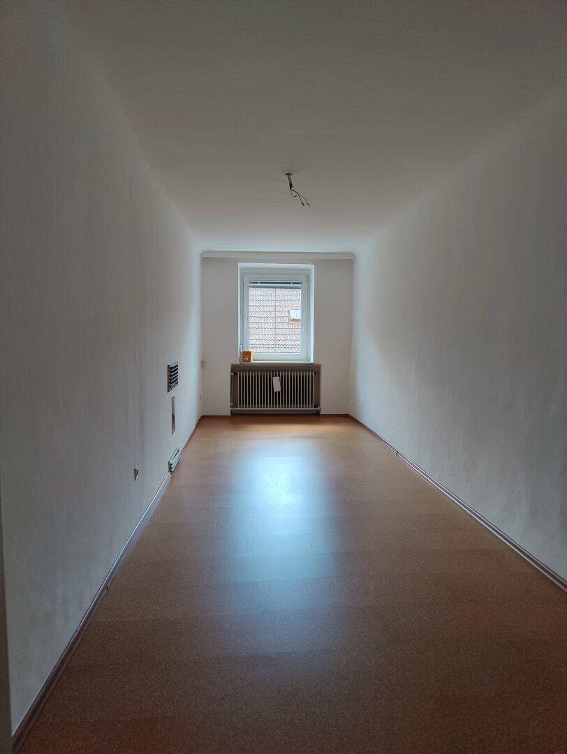 Langer, heller Raum mit Laminatboden und einem Fenster, das Tageslicht hereinlässt.