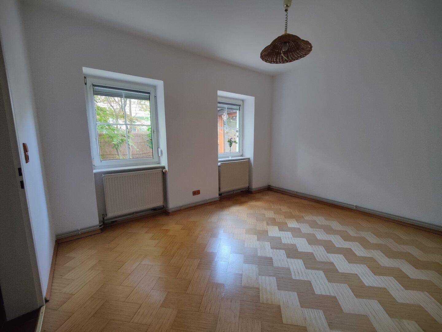 Heller Raum mit Fischgrätparkettboden und zwei Fenstern, die Tageslicht hereinlassen.
