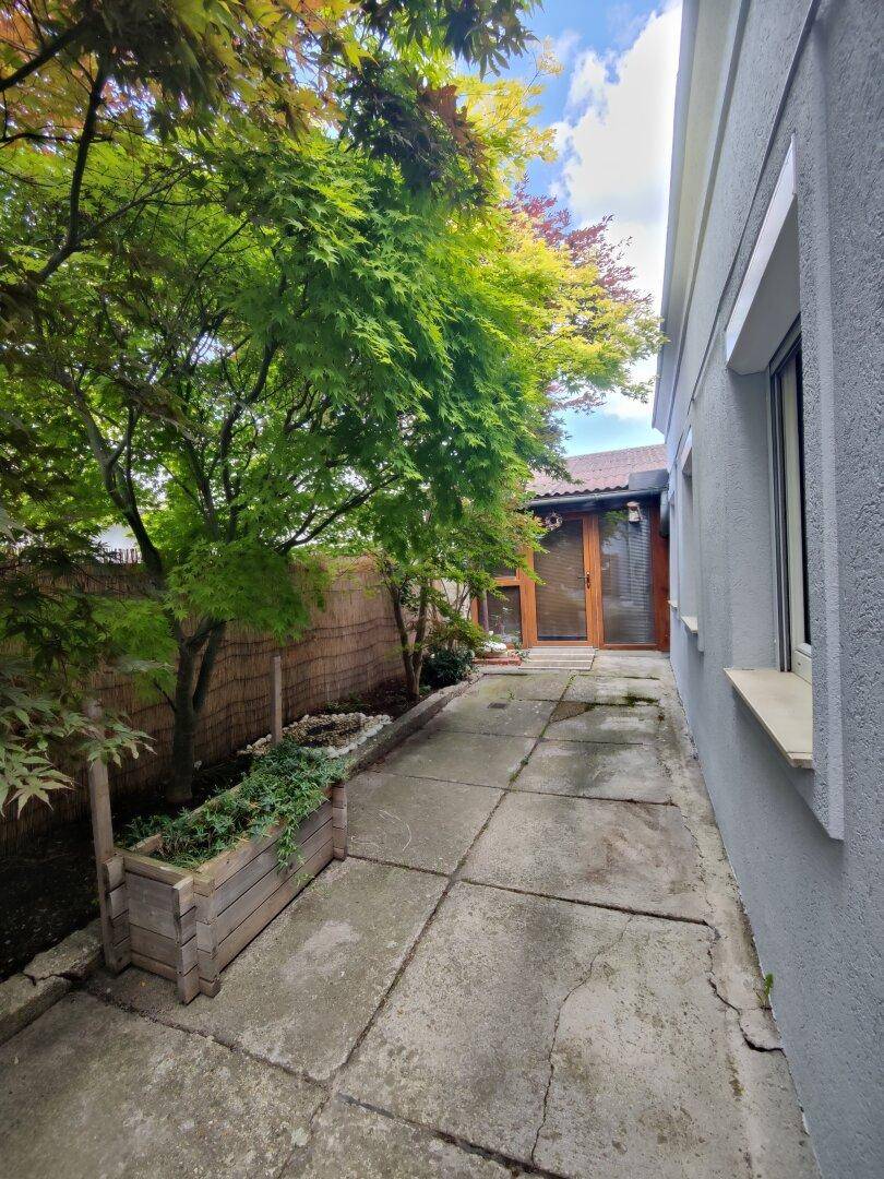 Schmaler gepflasterter Gartenweg entlang des Hauses mit Bäumen und Pflanzen.