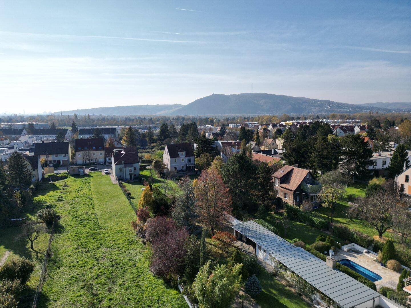Panoramablick aus der Vogelperspektive auf eine Wohngegend mit Häusern, Gärten, einem Pool und einem bewaldeten Hügel im Hintergrund.