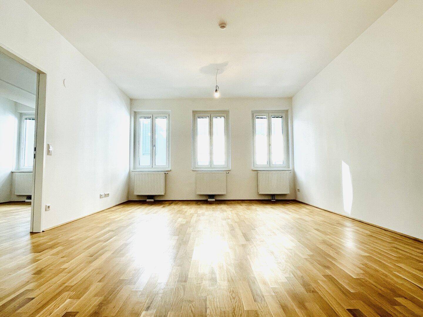 Helles, leeres Wohnzimmer mit Parkettboden und drei Fenstern, bereit zur Einrichtung.