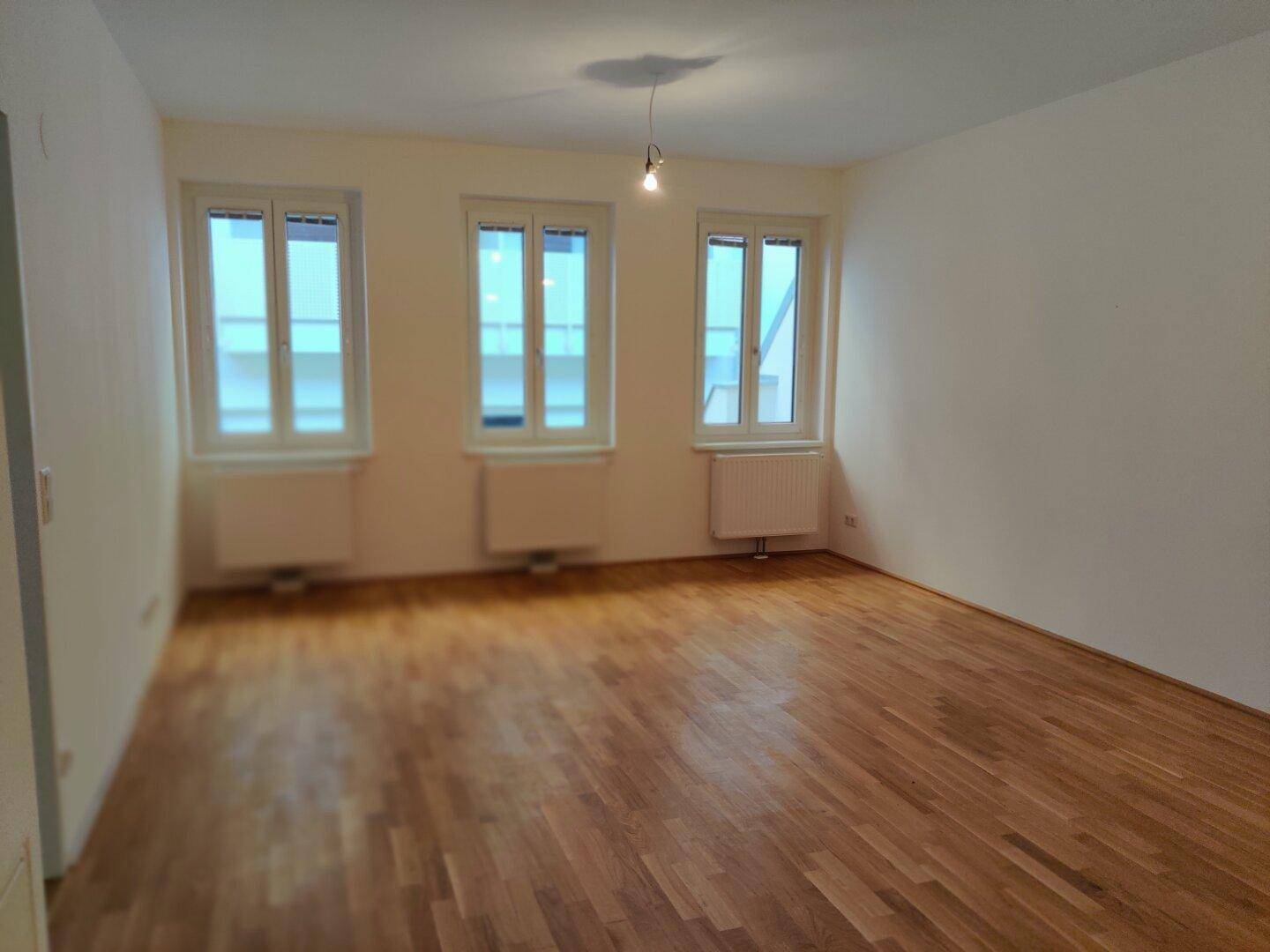 Lichtdurchflutetes Wohnzimmer mit Parkettboden, drei Fenstern und Heizkörpern unter den Fenstern.