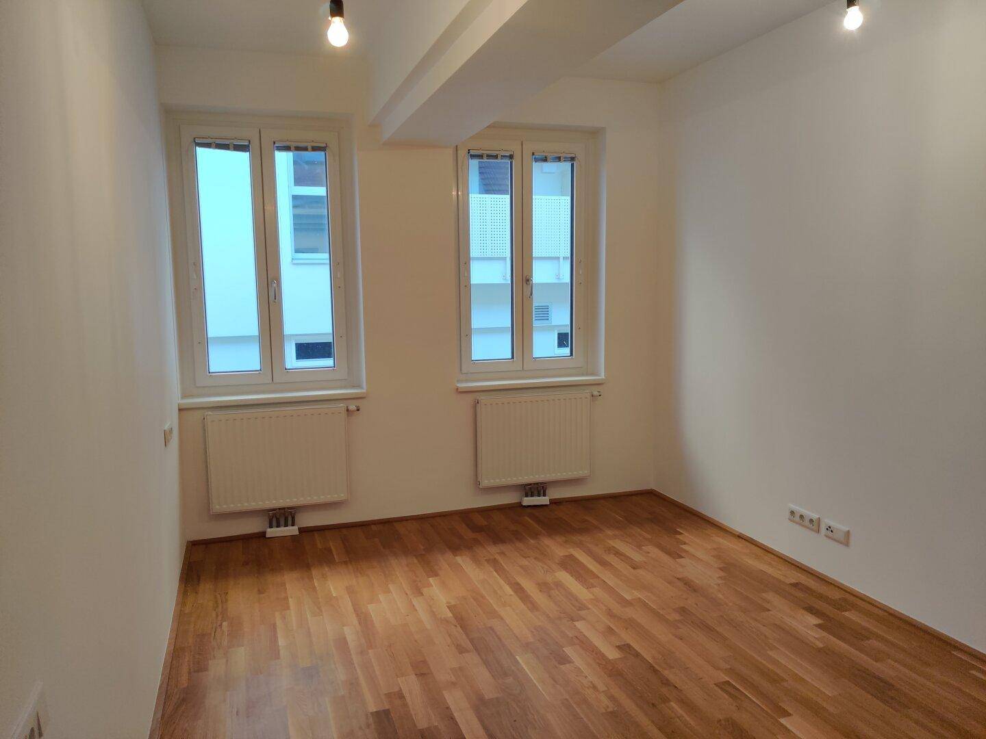 Helles Zimmer mit Parkettboden, zwei großen Fenstern und modernen Deckenleuchten.