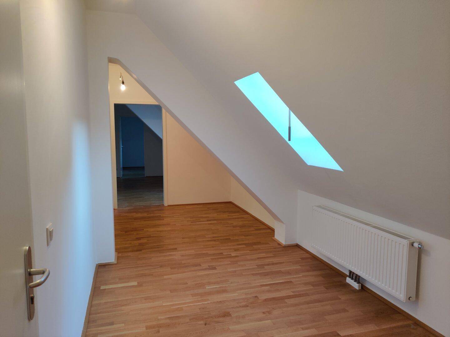 Dachgeschossraum mit schräger Decke, Parkettboden und einem Dachfenster, das Tageslicht spendet.