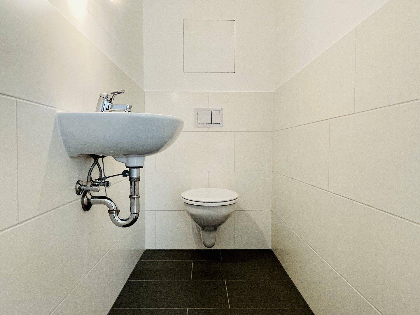 Gäste-WC mit wandhängender Toilette, kleinem Waschbecken und weißen Wandfliesen.