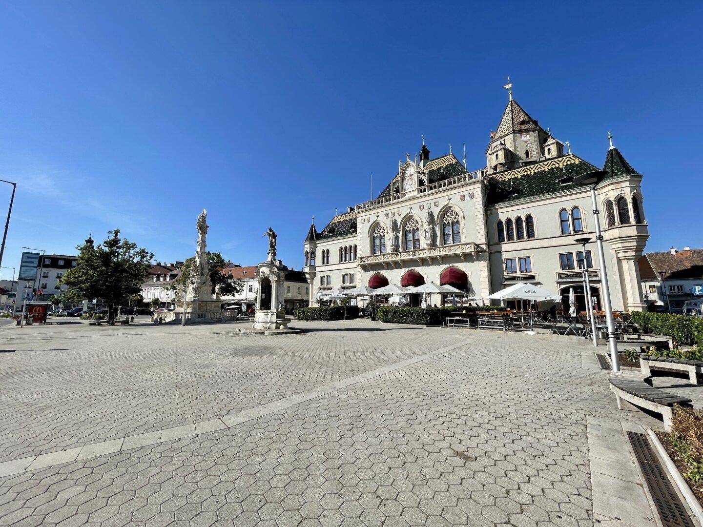 Historischer Stadtplatz mit imposantem Gebäude, Denkmal und Außengastronomie unter blauem Himmel.