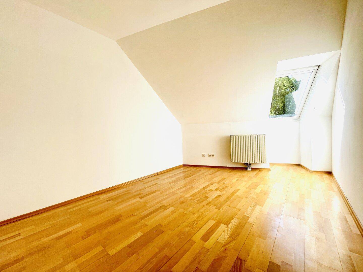 Helles Zimmer im Dachgeschoss mit Parkettboden, schräger Decke und Blick ins Grüne.