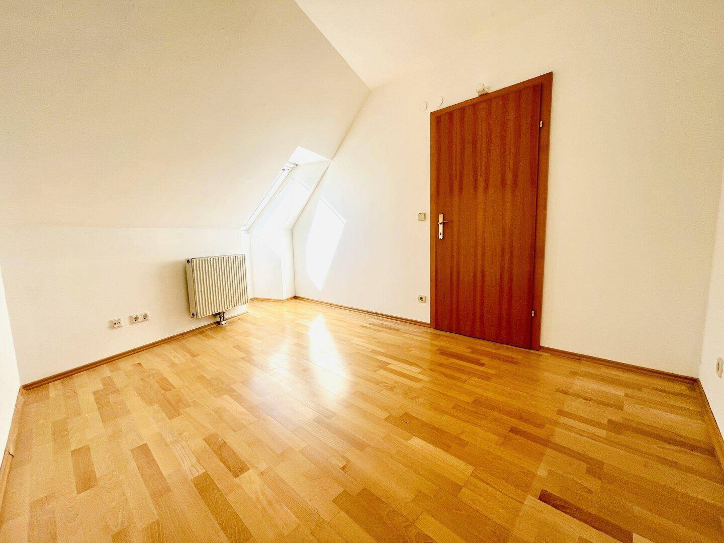 Leeres Zimmer im Dachgeschoss mit Parkettboden, schräger Decke und Heizkörper.