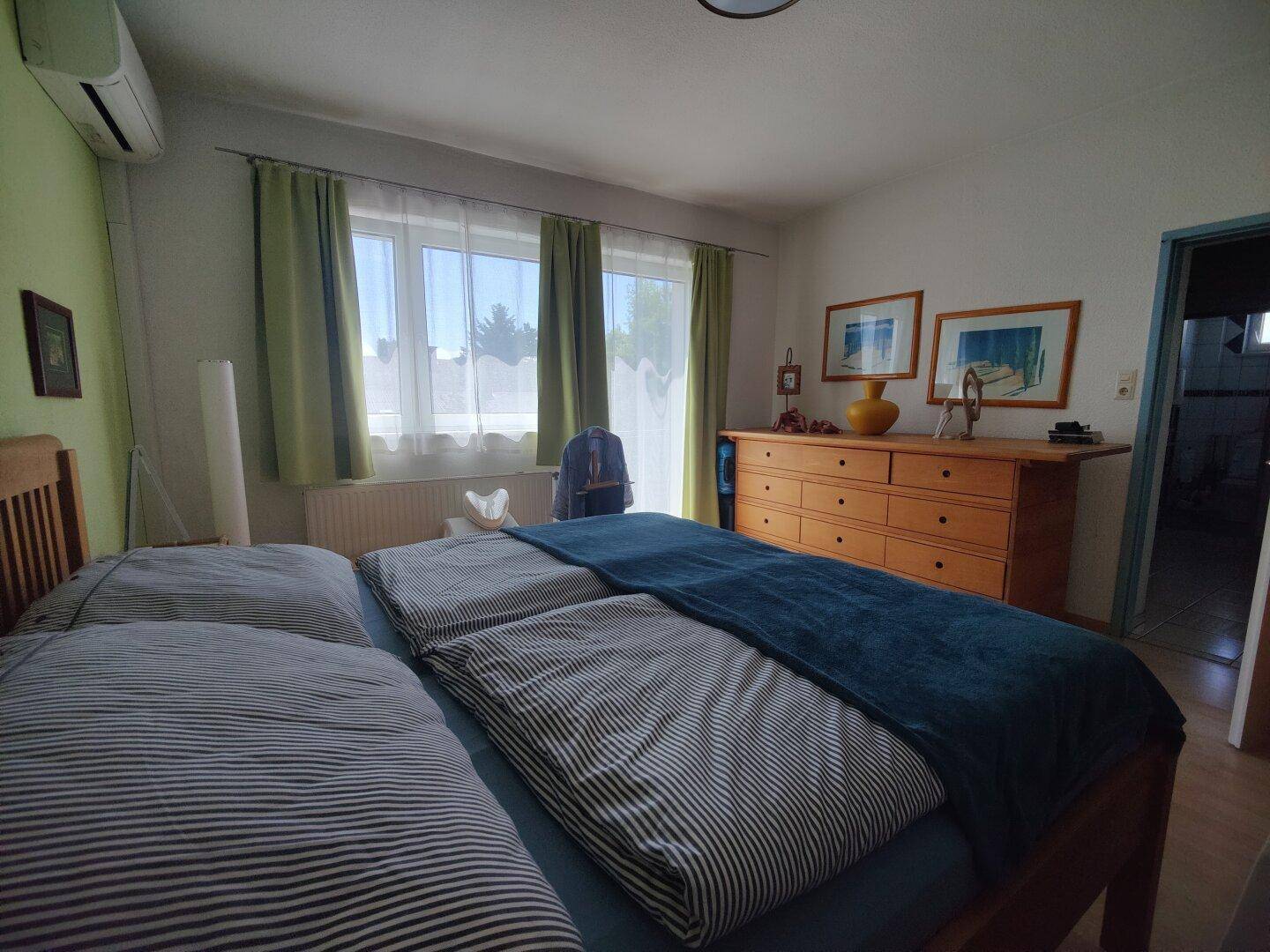 Helles Schlafzimmer mit Doppelbett, Holzkommode und Klimaanlage, mit Zugang zum Balkon.