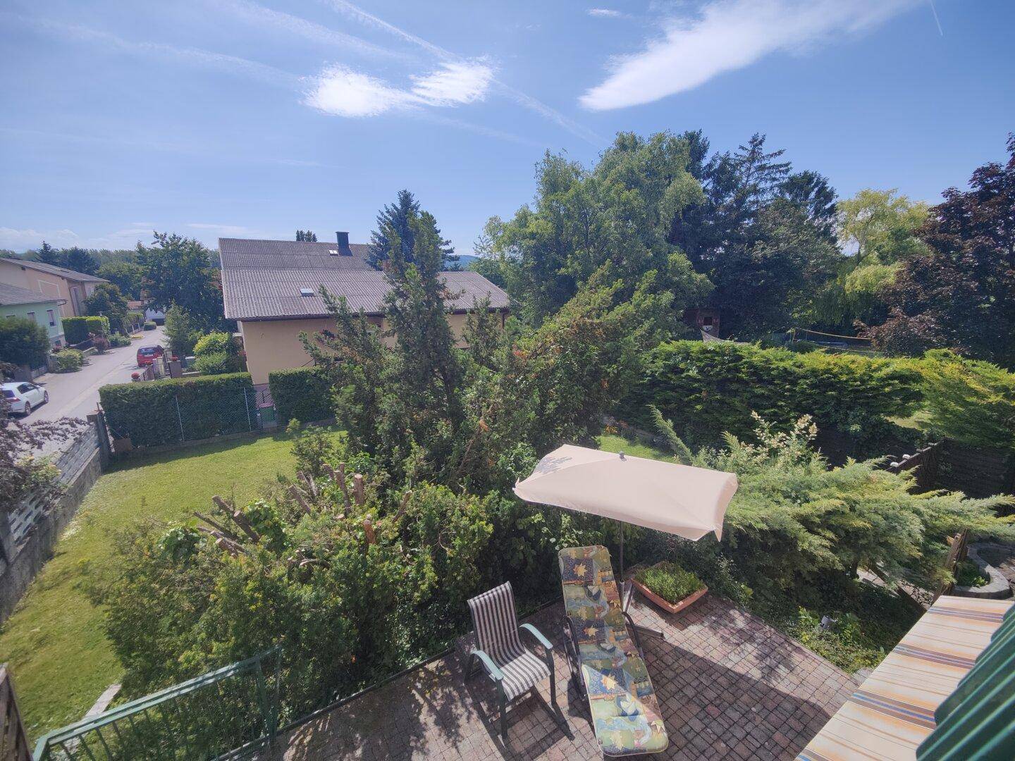 Sonniger Terrassenbereich mit Liegestuhl und Sonnenschirm, umgeben von viel Grün.