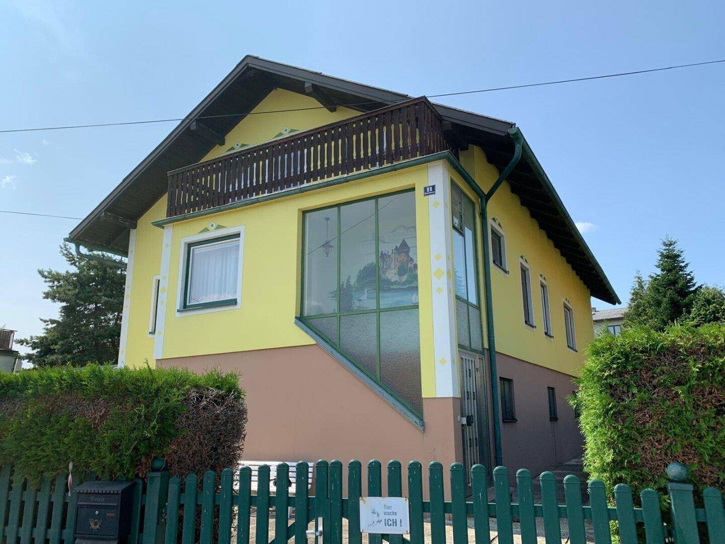 Vorderansicht des gelben Hauses mit Balkon, Wintergarten und grüner Hecke.