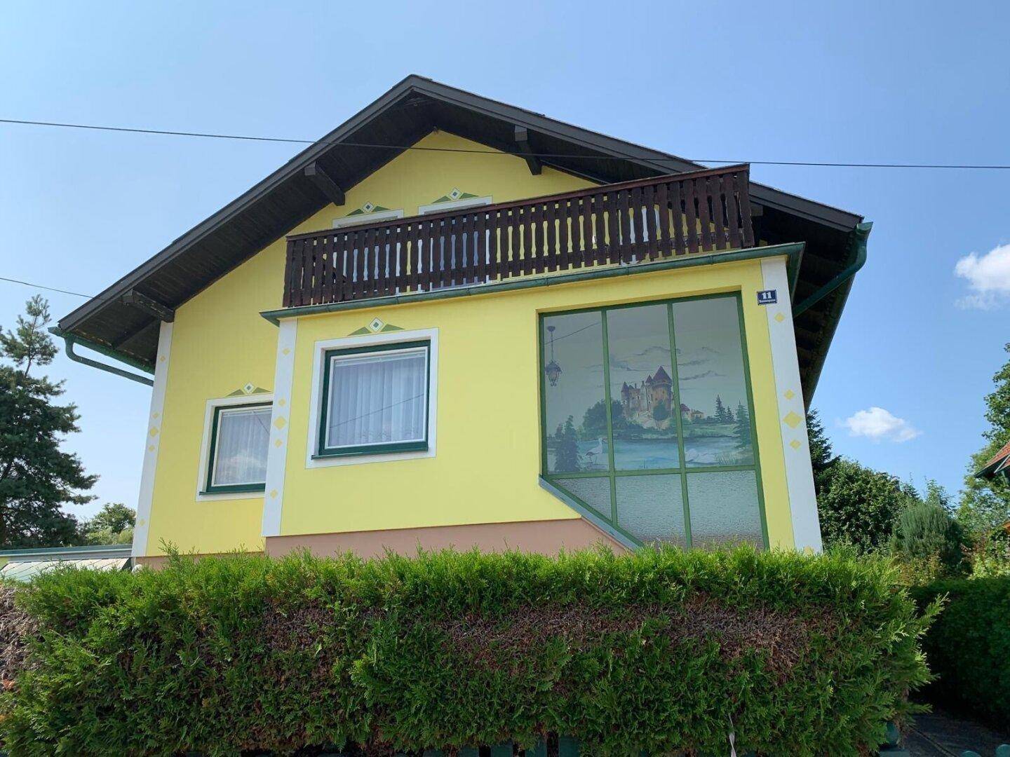 Gelbes Wohnhaus mit Balkon, verglastem Anbau und gepflegter Hecke im Vorgarten.