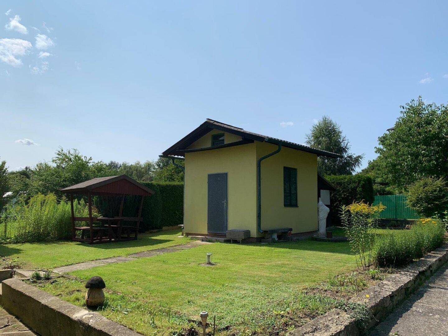 Garten mit kleinem Nebengebäude, Holzpavillon und Rasenfläche unter blauem Himmel.