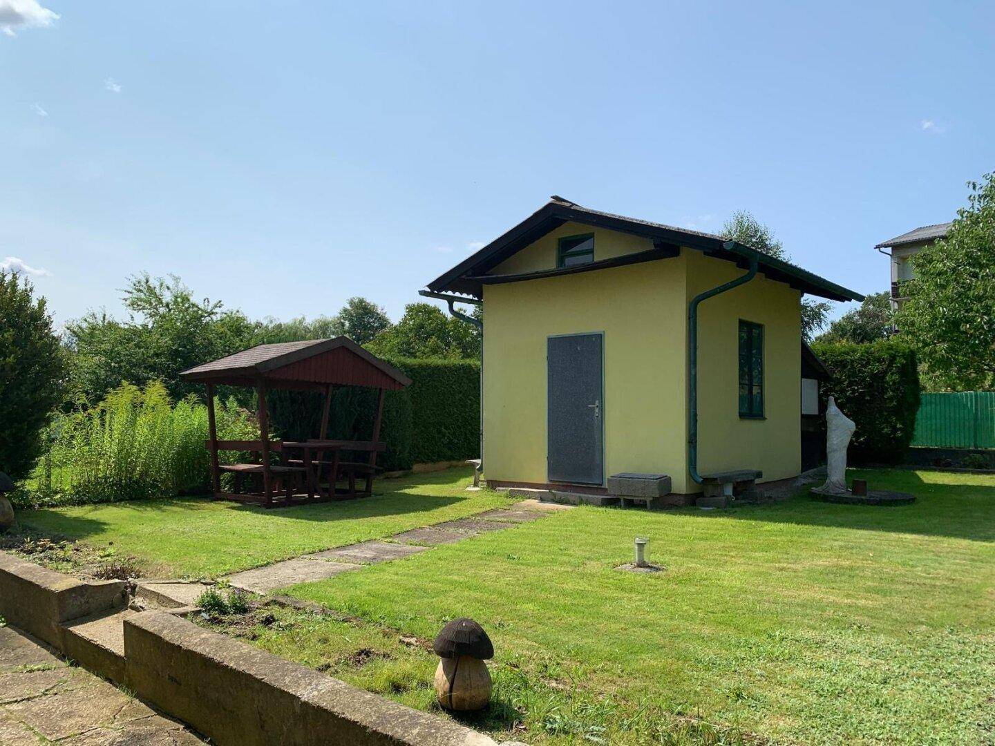 Gartenbereich mit kleinem Gartenhaus, Holzpavillon und gepflegtem Rasen an einem sonnigen Tag.