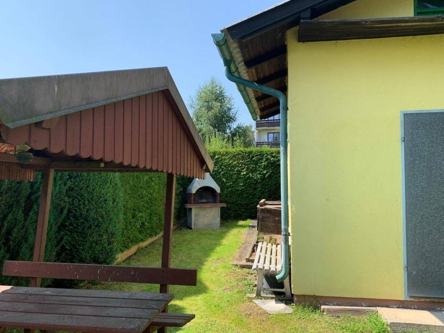 Gartenbereich mit gemauertem Grillplatz und Holzpavillon, umgeben von einer hohen Hecke.