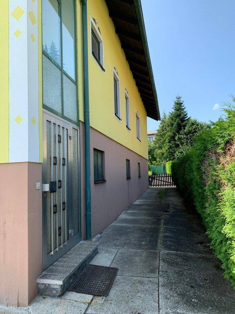 Seitenansicht des Hauses mit Eingangsbereich, schmalem Weg und hoher Hecke.