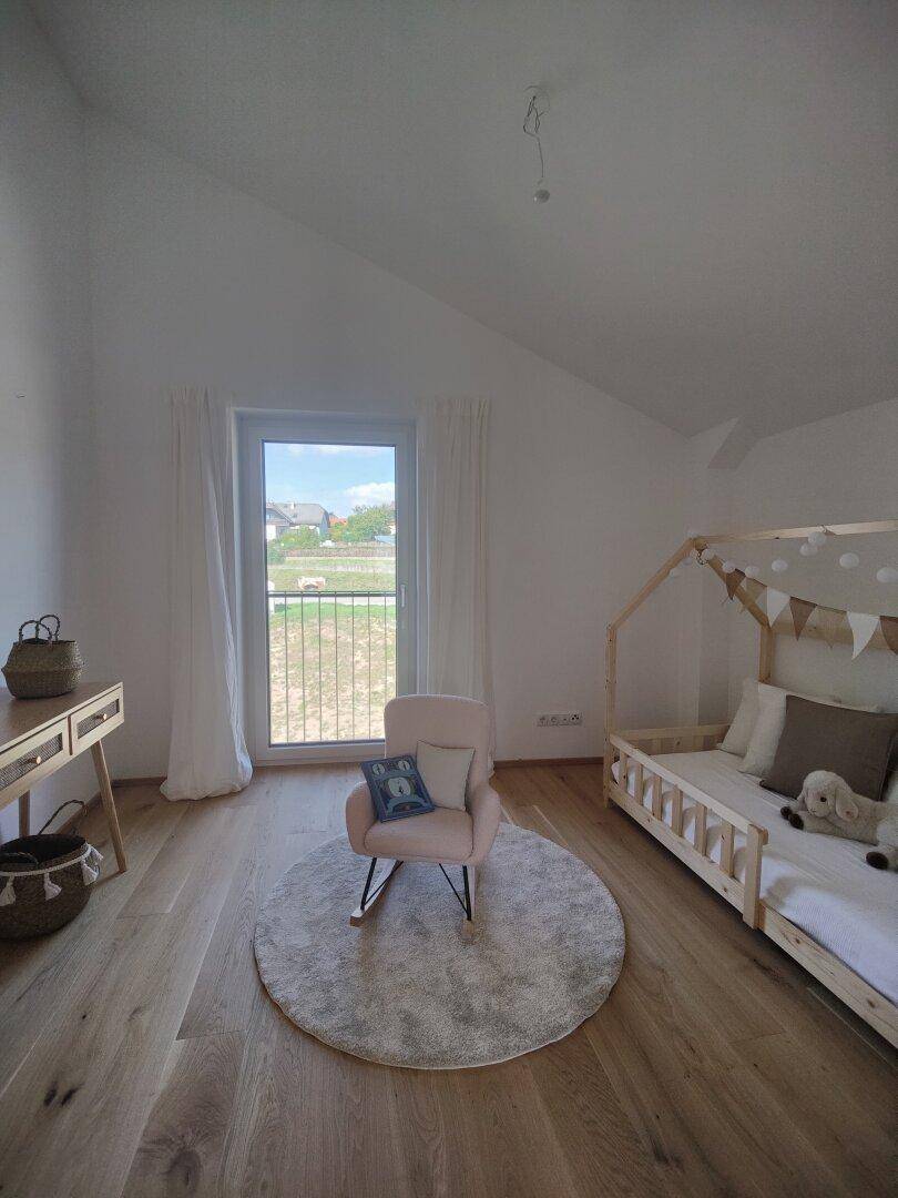 Freundliches Kinderzimmer mit Holzboden, Babybett und Schaukelstuhl am Fenster.