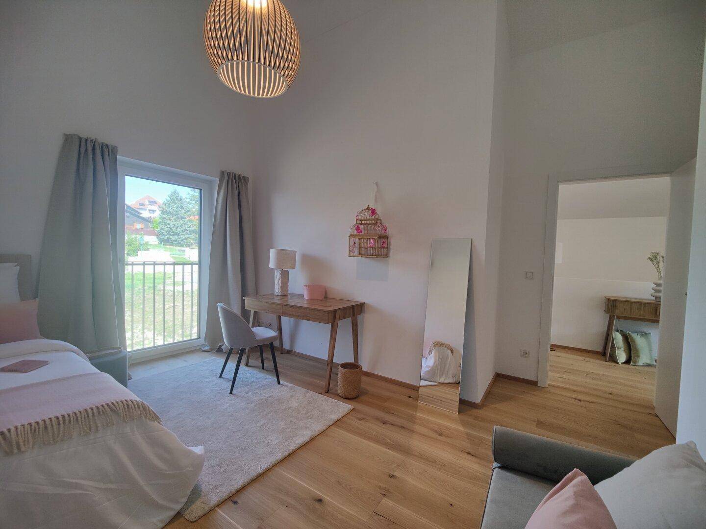 Helles Schlafzimmer mit Holzboden, Schreibtisch und Zugang zu einem Balkon.