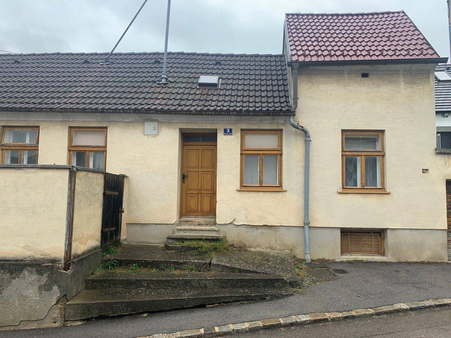 Vorderansicht eines älteren Hauses mit Holztür und Fenstern, direkt an einer leicht ansteigenden Straße gelegen.