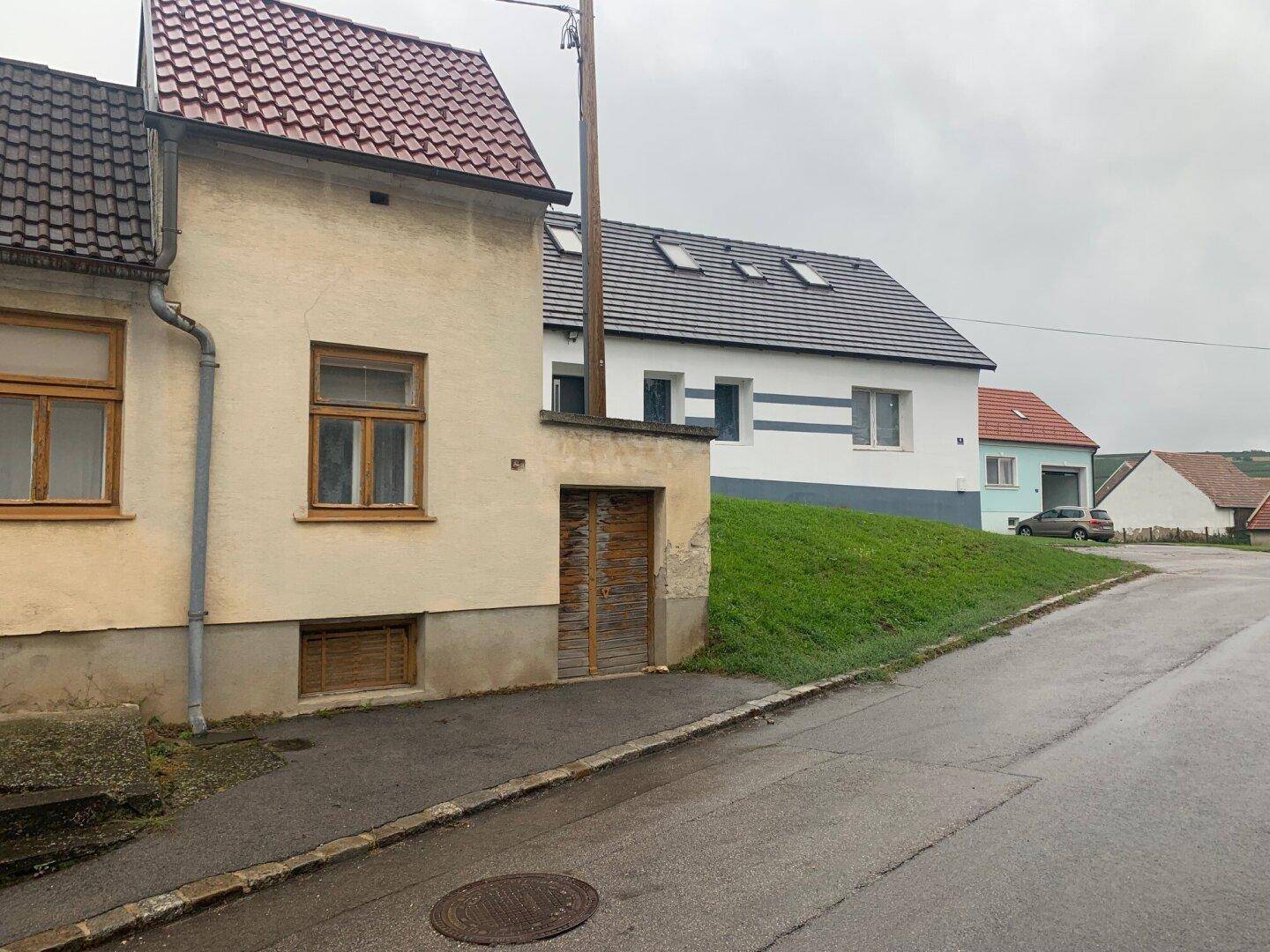 Blick auf die Straße mit dem älteren Haus im Vordergrund und einem Neubau auf einem grünen Hügel im Hintergrund.