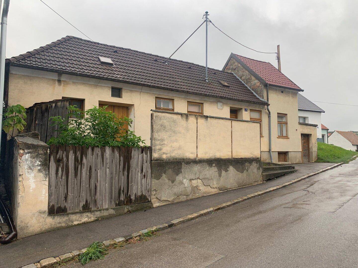 Seitenansicht eines älteren Hauses mit Putzfassade und Holzzaun an einer leicht ansteigenden Straße unter bewölktem Himmel.